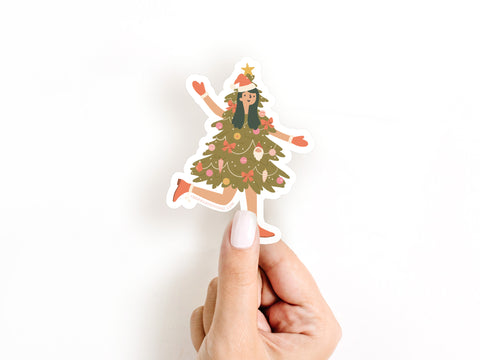 A hand with light skin holds Onderkast Studio’s Christmas Tree Girl Sticker, featuring a smiling person dressed as a decorated Christmas tree with ornaments, bows, and a star. This sticker is water resistant.