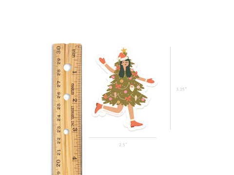 The Christmas Tree Girl Sticker by Onderkast Studio features a girl dressed as a festive tree with ornaments and a star. Made from durable, water-resistant vinyl, it measures about 2.5 wide by 3.25 tall. Shown beside a ruler for scale.