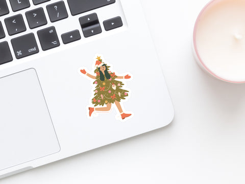 A laptop with the Christmas Tree Girl Sticker by Onderkast Studio, featuring a person dressed as a decorated Christmas tree, sits on a white surface next to a pink candle.