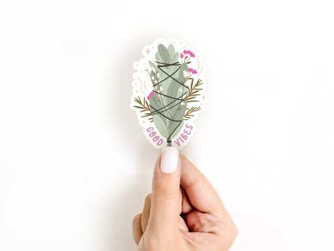 A hand with white nail polish holds the Onderkast Studio Good Vibes Smudge Stick Sticker, a durable vinyl decal featuring a sage bundle with string, small purple flowers, and “GOOD VIBES” on a white background.
