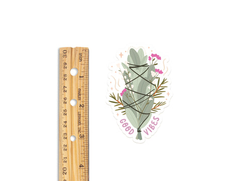 The Good Vibes Smudge Stick Sticker by Onderkast Studio features a durable, water-resistant vinyl design of a sage bundle with pink flowers, green leaves, black string, and GOOD VIBES below. Its about 3 inches tall.