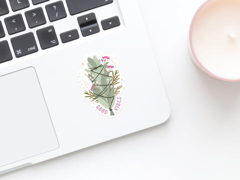A close-up of a laptop with an Onderkast Studio Good Vibes Smudge Stick Sticker next to the trackpad, and a lit candle in a pink holder on a white surface nearby.
