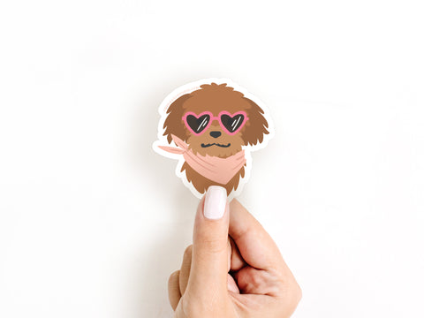 A hand with light-colored nail polish holds the Onderkast Studio Dog w/ Heart Sunglasses Sticker, a water-resistant vinyl sticker featuring a cartoon dog in heart-shaped sunglasses and a pink scarf, shown against a plain white background.