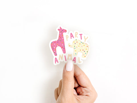 A hand with light pink nail polish holds Onderkast Studios Frosted Party Animal Cookies Sticker, featuring a playful pink giraffe and yellow polka-dot elephant with PARTY ANIMAL in vibrant, durable vinyl on a white background.