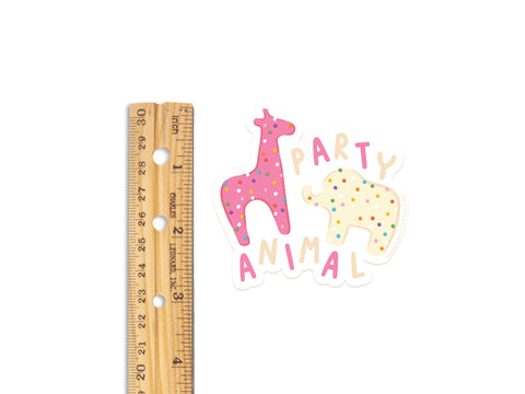 A wooden ruler next to Onderkast Studio’s Frosted Party Animal Cookies Sticker, a durable vinyl sticker with vibrant colors on white—featuring a pink polka dot giraffe, yellow polka dot elephant, and playful “PARTY ANIMAL” text.