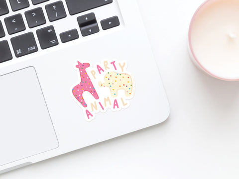 A laptop featuring Onderkast Studios Frosted Party Animal Cookies Sticker, with a pink giraffe and yellow elephant beside PARTY ANIMAL. A white candle in a pink holder appears on the right, enhancing the vibrant scene.