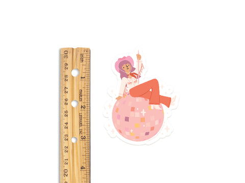 The Disco Cowgirl Sticker by Onderkast Studio is a durable, water-resistant vinyl sticker showing a person in a pink hat and orange boots sitting on a pink disco ball. Measures approx. 2.5 tall (ruler for scale).