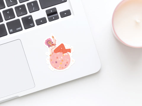 A laptop featuring the Onderkast Studio Disco Cowgirl Sticker, showing a cartoon character in an orange outfit lying on a pink disco ball. A white candle in a pink holder sits nearby on a white surface.