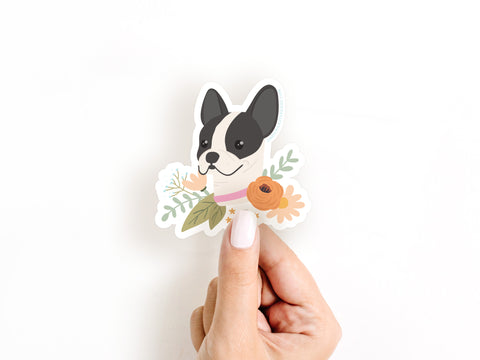 A hand with white-painted nails holds the Onderkast Studio French Bulldog Floral Sticker, a water and sunlight resistant vinyl featuring a black and white French bulldog with illustrated orange and yellow flowers on a white background.