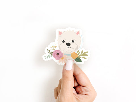 A hand with white painted nails holds the Onderkast Studio White Terrier Floral Sticker, a cute and durable vinyl sticker featuring a white dog with flowers around its neck, against a plain white background.