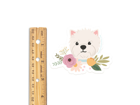 The Onderkast Studio White Terrier Floral Sticker features a cute, fluffy white dog with colorful flowers around its neck. This durable, water-resistant vinyl sticker measures about 2 inches tall and is shown next to a ruler for scale.