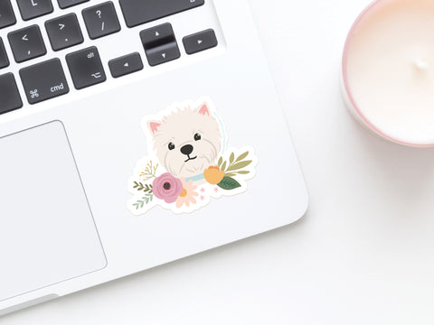 A white laptop features the Onderkast Studio White Terrier Floral Sticker, a cute and durable vinyl sticker of a white dog with colorful flowers, displayed beside a pink candle.