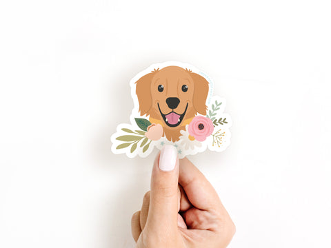 A hand with light pink nail polish holds the Onderkast Studio Golden Retriever Floral Sticker, a water-resistant sticker featuring a smiling cartoon golden retriever among flowers and leaves, shown on a plain white background.