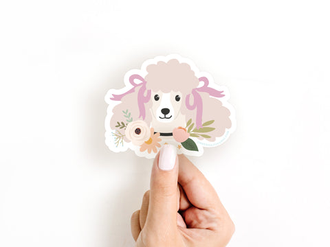 A manicured hand holds the Onderkast Studio Poodle Floral Sticker—a durable vinyl sticker featuring a light pink poodle with bows, surrounded by vibrant flowers and leaves on a white background.