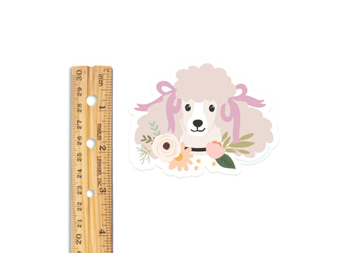A wooden ruler lies next to Onderkast Studios Poodle Floral Sticker, a vibrant, durable vinyl sticker featuring a cream poodle with pink bows and flowers around its neck, all set against a white background.