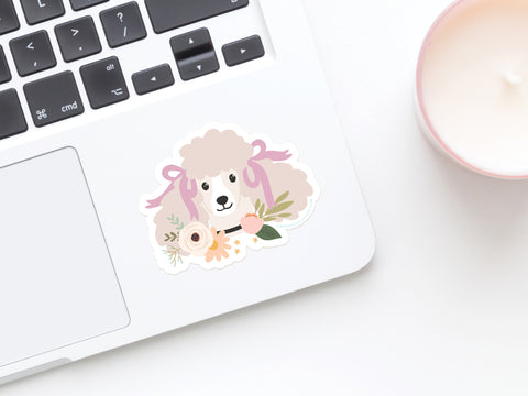 The Onderkast Studio Poodle Floral Sticker decorates a laptop palm rest, showing a poodle with pastel flowers. A pale pink candle in a glass jar sits nearby on the white surface.
