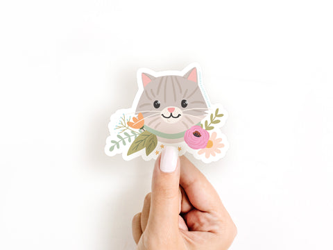 A hand with light-colored nail polish holds the Tabby Cat Floral Sticker by Onderkast Studio, featuring a smiling gray cat with a green collar, surrounded by colorful flowers and leaves, against a white background. Water resistant.