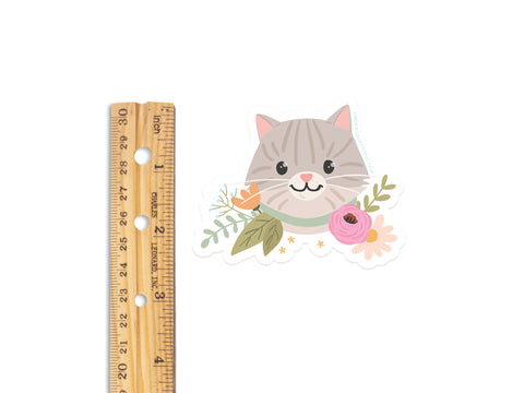 The Onderkast Studio Tabby Cat Floral Sticker—a cute, durable, water-resistant vinyl sticker featuring a gray tabby with big eyes and a pink nose—measures about 3 inches wide and has charming flowers around its neck.