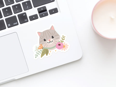 The Tabby Cat Floral Sticker by Onderkast Studio, a cute and water-resistant sticker featuring a gray tabby cat with flowers, is shown on a white laptop next to a pink cup holding a candle.