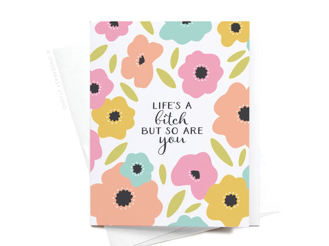 The Life’s a Bitch But So Are You Greeting Card by Onderkast Studio features colorful stylized flowers and the bold phrase “Life’s a bitch but so are you,” with a partially visible envelope underneath.