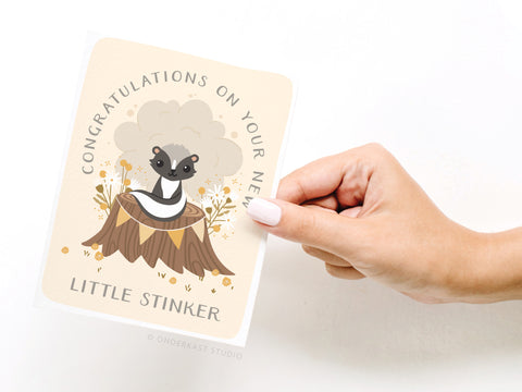 A hand holds Onderkast Studios Congratulations On Your New Little Stinker Greeting Card, featuring a cartoon skunk on a flower-surrounded tree stump—perfect for congratulating new parents.