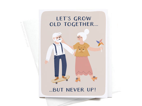 The Let’s Grow Old Together Greeting Card by Onderkast Studio features an elderly man skateboarding, holding hands with an elderly woman with a pinwheel. The light-colored card reads, “LET’S GROW OLD TOGETHER… BUT NEVER UP!”.