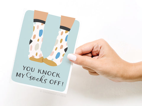 A hand holds Onderkast Studio’s You Knock My Socks Off Greeting Card – FINAL STOCK, featuring illustrated feet in polka-dot socks and the phrase You knock my socks off! on a light blue background.