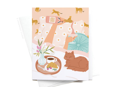 Cozy illustration of a person in bed with a phone, a napping cat, and breakfast—plus a daisy and cheetah blanket. Perfect for birthdays or funny notes. Rise & Wine Greeting Card – FINAL STOCK by Onderkast Studio.