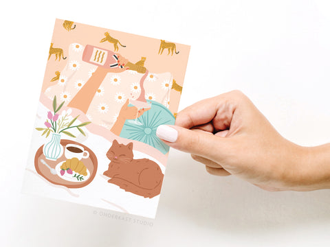 A hand holds the Rise & Wine Greeting Card – FINAL STOCK by Onderkast Studio, featuring a cozy illustration of a cat napping by breakfast, flowers in a vase, and legs on a leopard and daisy print blanket.