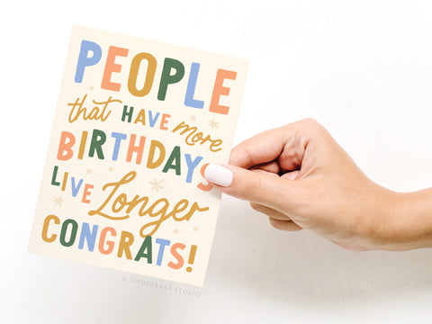 A hand with white-painted nails holds the Onderkast Studio “People That Have More Birthdays Live Longer Greeting Card,” featuring colorful, playful lettering—printed in the USA.