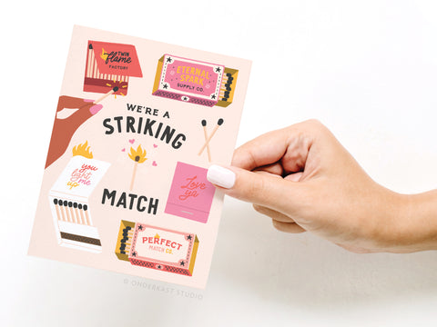 A hand holds Onderkast Studios Were a Striking Match Greeting Card, decorated with illustrated matchboxes, matches, and sweet messages like “We’re a striking match,” “You light me up,” and “Perfect match co.”.