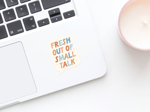 A laptop with the Onderkast Studio Fresh Out Of Small Talk Sticker by the keyboard—ideal for introverts. Nearby, a white candle in a pink holder rests on a white surface, adding a cozy touch.
